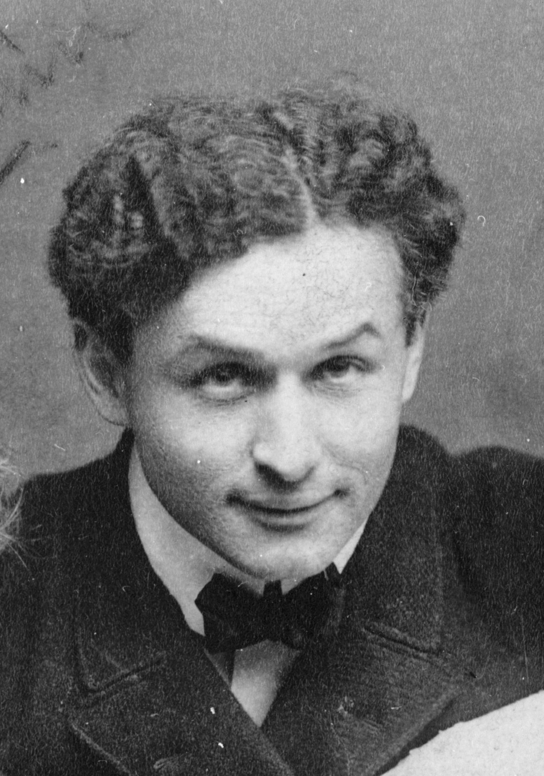 Close-up black and white portrait of a young Harry Houdini with curly hair, a dark coat and a bow tie, smiling slightly