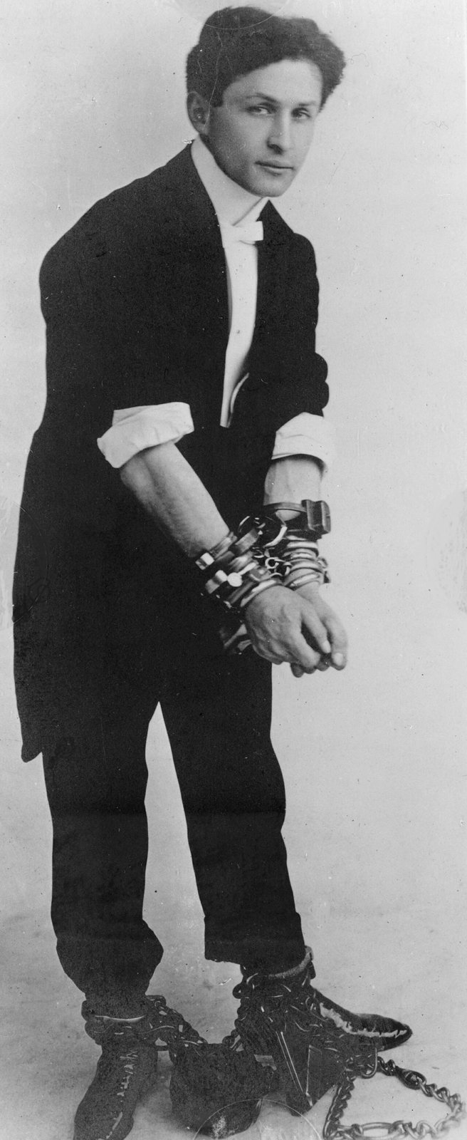 Harry Houdini in a dark suit with his wrists locked in several sets of handcuffs and his ankles chained (Photo: unknown author, public domain, via Wikimedia Commons)