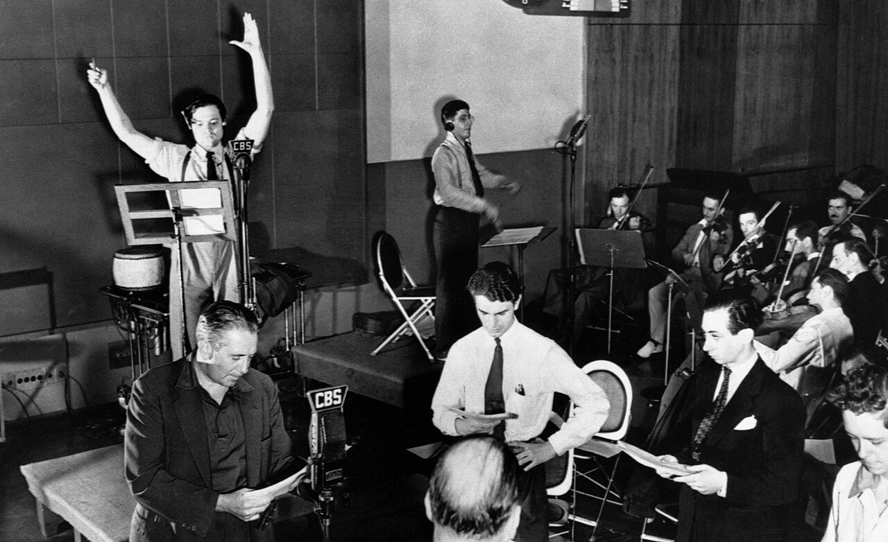 Orson Welles, arms raised, conducting a Mercury Theatre radio rehearsal at CBS in 1938 with actors and an orchestra (Photo: Acme Telephoto, public domain, via Wikimedia Commons)