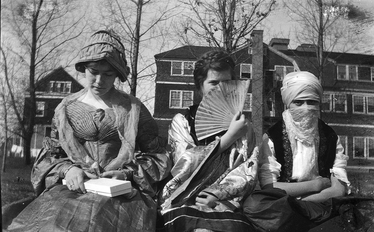 Three young women in homemade costumes sit on a lawn in 1918, one in a bonnet, one hiding behind a fan, one in a turban and veil