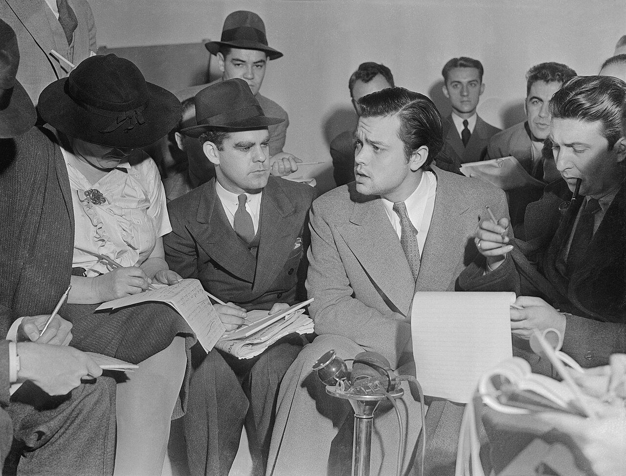 Orson Welles in a light suit talking to a crowd of reporters with notepads, a microphone stand in front of him
